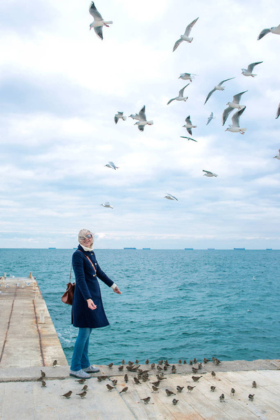 mulher loira alimentando gaivotas no dia nublado de outono na costa do mar
  - Foto, Imagem