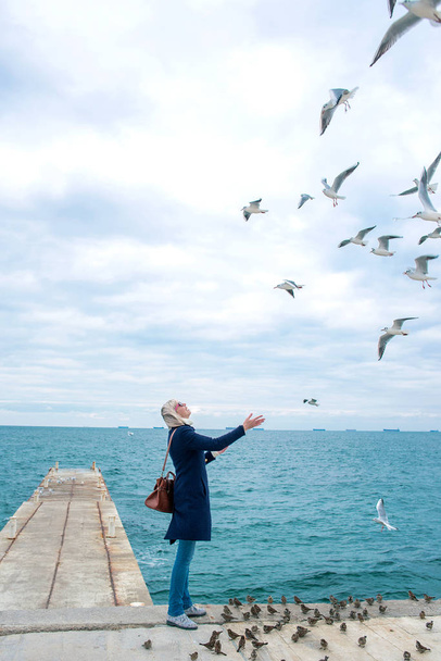 blonde woman feeding seagulls in cloudy autumn day on the sea coast  - Photo, Image