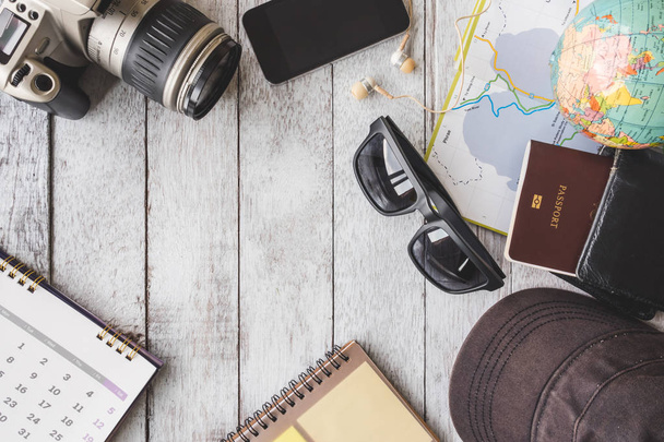 Το Top view κάμερα ημερολόγιο, έξυπνο τηλέφωνο, ακουστικά, γυαλιά ηλίου, καπάκι, σημειωματάριο, πορτοφόλι, διαβατήριο, Χάρτης και υδρόγειο σε φόντο λευκό ξύλινο τραπέζι, έννοια του ταξιδιού - Φωτογραφία, εικόνα