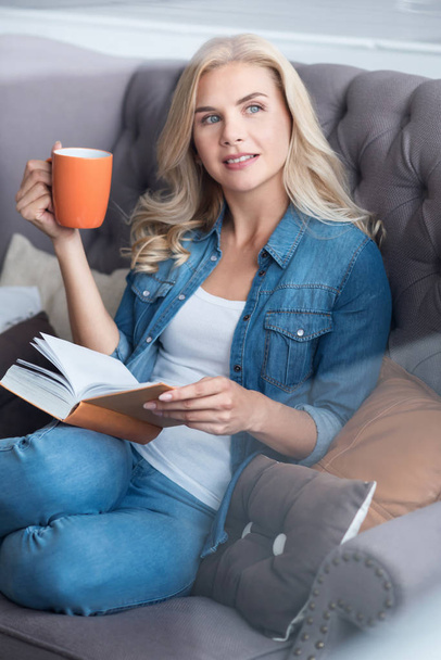 junge blonde Frau sitzt auf Couch mit Buch und Tasse - Foto, Bild
