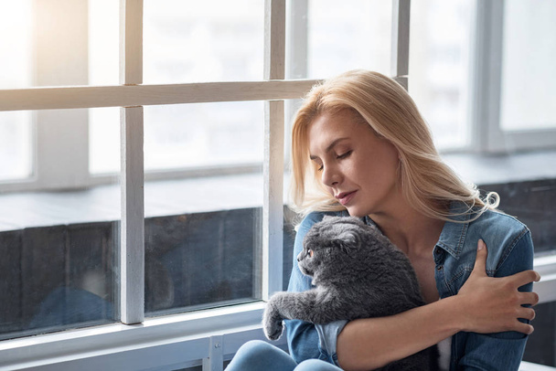 Blond woman holding cat near window. - Foto, immagini