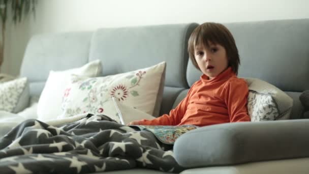 Little boy, reading a magazine, sitting on a sofa, covered with soft blanket - Imágenes, Vídeo
