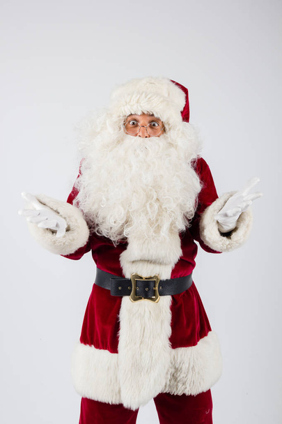 Санта-Клаус в очках и красном костюме бросает руки на белом фоне и смотрит в камеру
.  - Фото, изображение