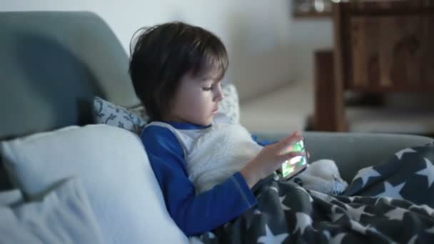 Little preschool child, boy, playing on mobile phone on the sofa at night - Materiał filmowy, wideo