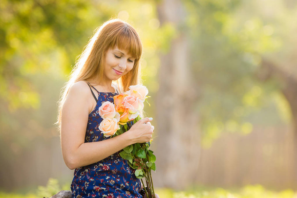 Красивая молодая сексуальная девушка смотрит на букет роз, подаренных ей стоя на фоне зеленого солнечного размытого
 - Фото, изображение