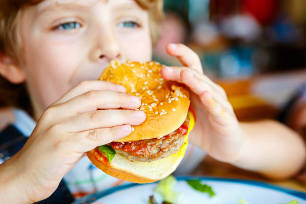 Cute healthy preschool boy eats hamburger sitting in cafe outdoors - Photo, Image