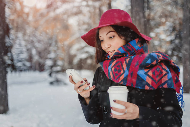 Μοντέρνα ντυμένος γυναίκα σε ένα πανωφόρι και πλατύγυρο καπέλο στο χειμερινό πάρκο πίνοντας καφέ και μιλάμε για το smartphone - Φωτογραφία, εικόνα