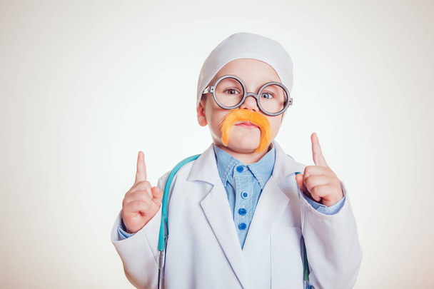 Happy little boy in doctor costum with glasses and mustache stand holding up two fingers on white background. - Fotografie, Obrázek
