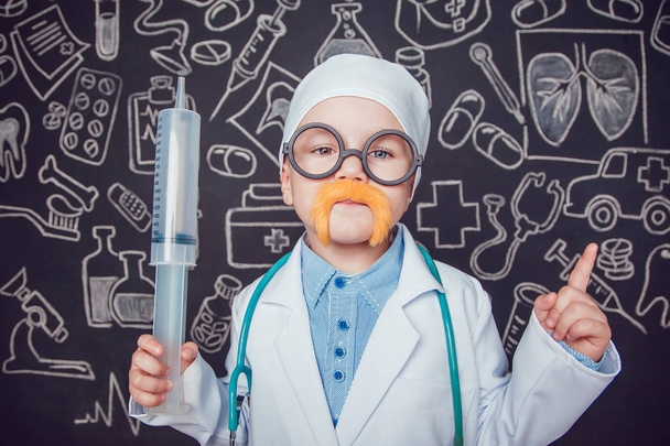 Happy little boy in doctor costume holding syringe on dark background with pattern. The child has mustache and glasses. Lifts thumbs up. - Photo, image