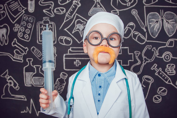 Happy little boy in doctor costume holding syringe on dark background with pattern. The child has mustache and glasses. - Photo, Image