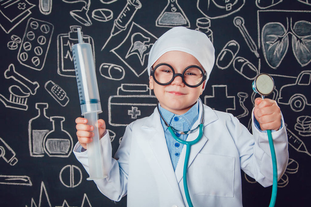 Happy little boy in doctor costum holding syringe and sthetoscope on dark background with pattern. The child has glasses - Foto, imagen