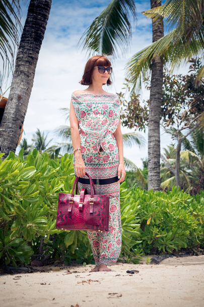Sexy bruneta modelka pózuje na pláži. Nosí brýle a luxusní červené handmande hadí kůže python kabelky. Bali, Indonésie. - Fotografie, Obrázek