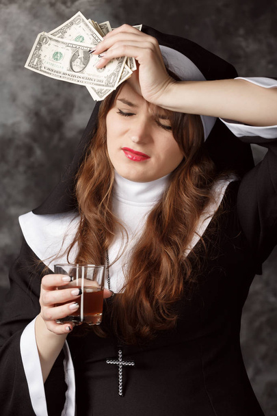 Portret van een nun holding dollarbiljetten en een glas met alcohol op een donkere achtergrond - Foto, afbeelding