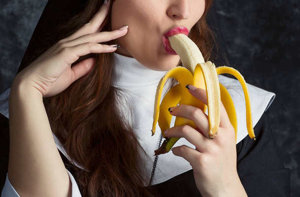 ελκυστική μοναχή, κρατώντας μια μπανάνα και δαγκώνει τον σε σκούρο φόντο - Φωτογραφία, εικόνα