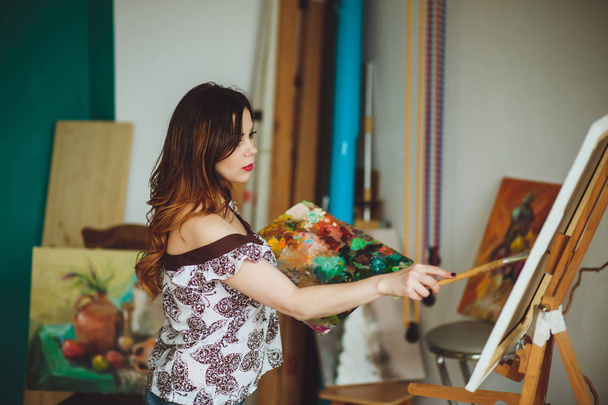 Woman artist painting a picture in a studio. Creative pensive painter girl paints a colorful picture on canvas with oil colors in workshop. - Photo, Image