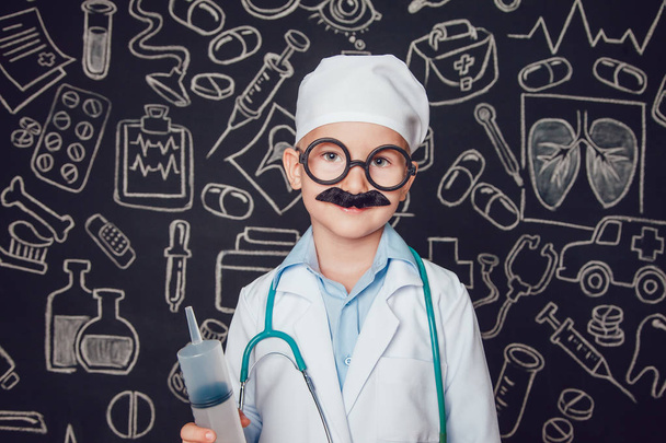 Little boy in doctor costume holding syringe on dark background with pattern. The child has mustache and glasses - Photo, image