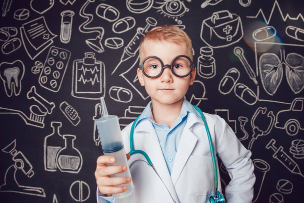 Happy little boy in doctor costume holding syringe on dark background with pattern. The child has glasses - Foto, imagen