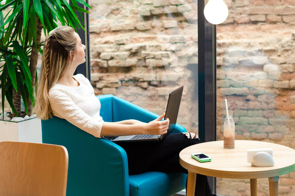 Portré, szép mosolygó nő ül egy kényelmes székbe, fekete laptop a kávézóban. Szép diák munka laptop. - Fotó, kép