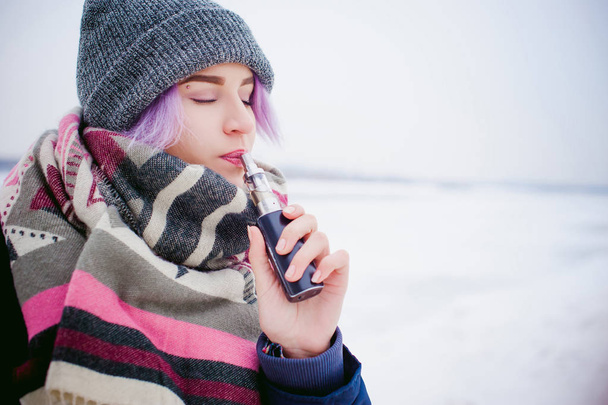 vaping girl. winter street portrait of a woman hipster, purple-dyed hair, a gray knitted hat and scarf. woman smokes an electronic cigarette in the street near the snow-covered river - Фото, зображення