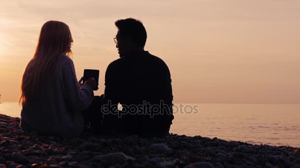 Technology in nature. Asian man wearing glasses and a woman have a rest on the nature, enjoy the tablet. At sunset near the sea or lake - Footage, Video