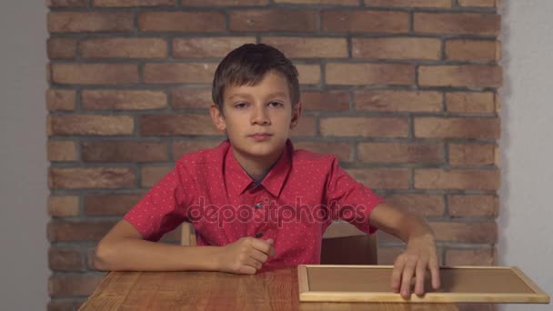 Παιδί κάθεται στο γραφείο κρατώντας πινακάκι με γράμματα Νορ στον τοίχο κόκκινο τούβλο φόντο. - Πλάνα, βίντεο