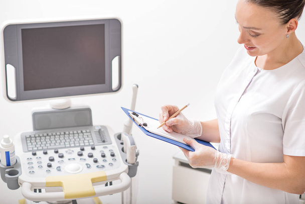 Cosmetologist making notes near ultrasound device - Zdjęcie, obraz