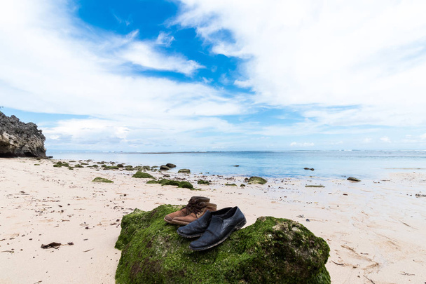 Old shoes on the big stone with moss. Small rocks scattered on beach sand close up. Beautiful ocean landscape, amazing sky. Bali, Indonesia. - Fotografie, Obrázek