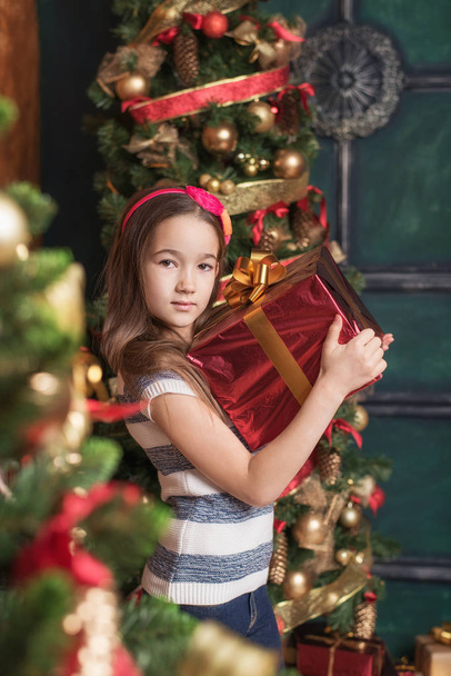 Cute little girl wearing red headband holding gift near christmas tree. - Fotografie, Obrázek