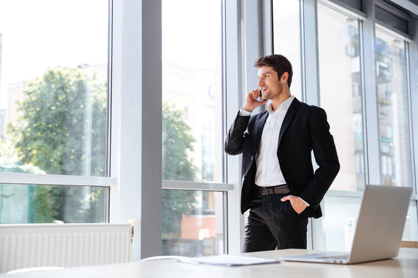 Smiling businessman standing and talking on cell phone in office - Foto, Bild