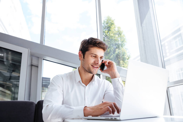 Happy young businessman talking on cell phone and using laptop - Foto, Imagen