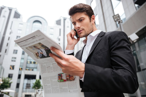 Concentrated young businessman talking on cell phone and reading newspaper - Foto, afbeelding
