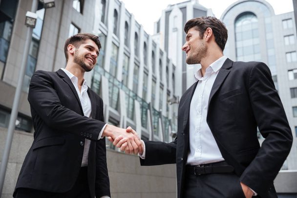 Two happy businessmen standing and shaking hands near business center - Photo, image