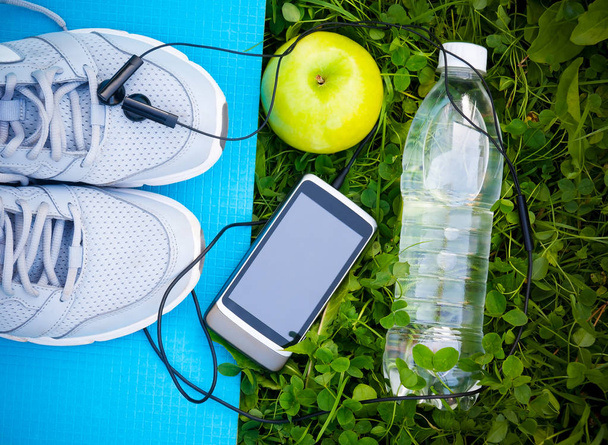 Sports shoes sneakers on yoga mat  and smartphone with headphones and apple on fresh green grass.  - Photo, Image