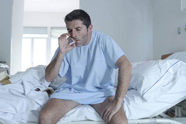 joven atractivo hombre buscando triste y preocupado en cama de hospital fumar cigarrillo en la clínica dormitorio
 - Foto, imagen