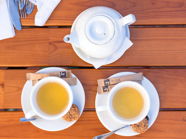 Duas xícaras brancas de chá e colheres com biscoitos, bule em uma mesa de madeira
 - Foto, Imagem