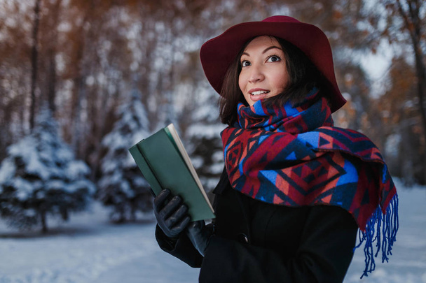 Beautiful young brunette woman reading book wearing knitted poncho, wide hat and coat in the snowy cold and magic winter forest. - Photo, Image