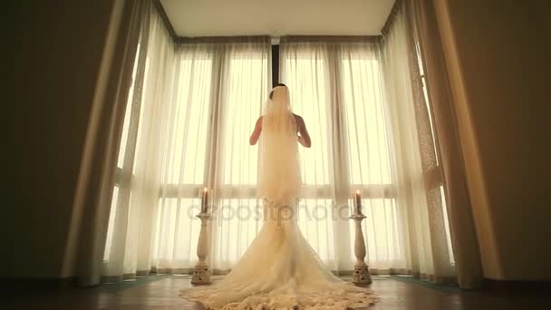 Zadní strana nevěsta ve svatebních šatech přijde do okna a odhalit bílé záclony. Teplé odstíny. - Záběry, video