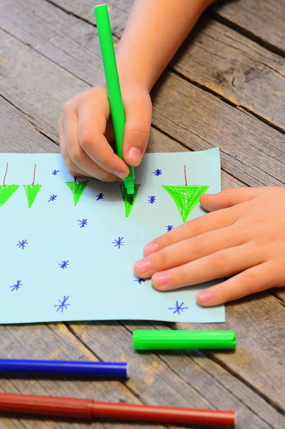 Malé dítě kreslí na papír vánoční stromky a sněhové vločky. Dítě v ruce drží zelená značka a kreslení stromu. Děti zimní obrázek, značky na dřevěný stůl. Předškolní výkresu aktivita - Fotografie, Obrázek