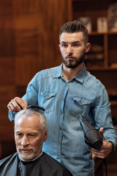 Handsome bearded hairdresser blow drying hair of client - Photo, image