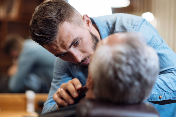 Attractive barber shaving beard of senior man - Фото, зображення