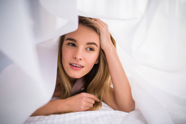 Smiling woman under a duvet in her bedroom. Closeup portrait of a beautiful young woman with blonde hair and under the blanket. happy good morning - Photo, Image