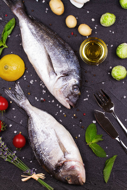 Raw fresh dorado fish with brussels sprouts, tomatoes, lemon, young potato, greens, bread, white wine bottle and olive oil on dark background - Foto, Imagen