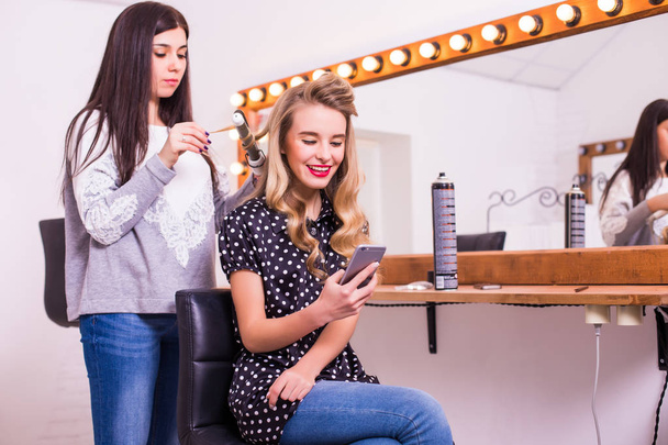 Cabeleireiro feminino aplicando alisador de cabelo para cabelos longos de jovem sorridente usando smartphone no camarim
 - Foto, Imagem