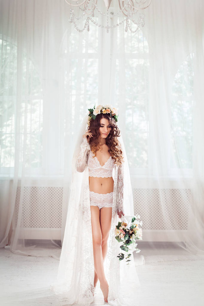 mariée sexy debout sur un hall de lumière blanche habillée en chemise de nuit, voile nuptiale et couronne, avec bouquet de fleurs
 - Photo, image