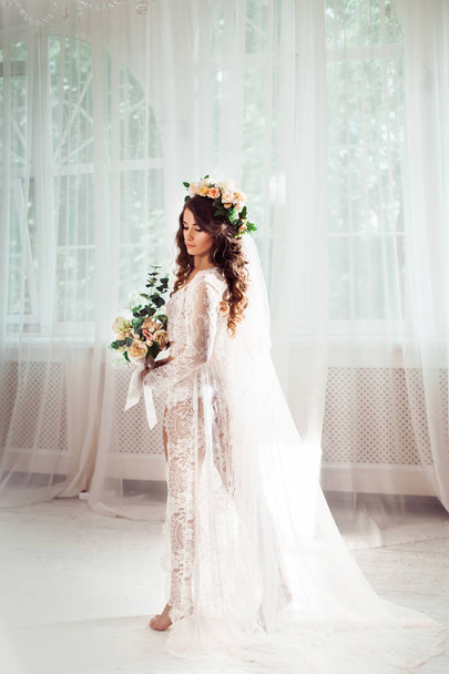 sexy novia de pie en un salón de luz blanca vestida con camisón, velo nupcial y corona, con ramo de flores
 - Foto, imagen