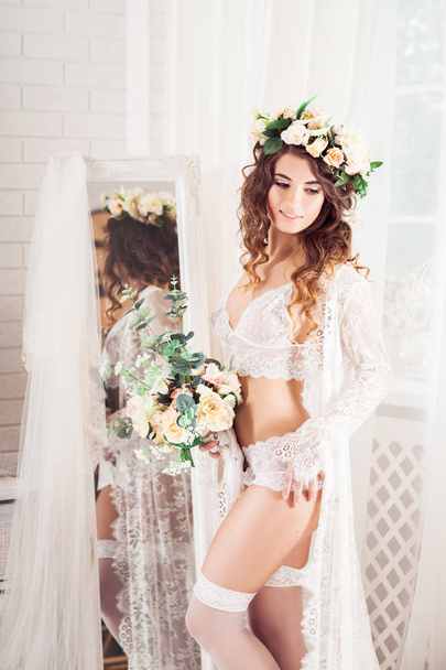 sexy caucasian bride standing in front of the full-length mirror, dressed in white nightie, wreath, with bouquet of flowers, vertical - Foto, imagen
