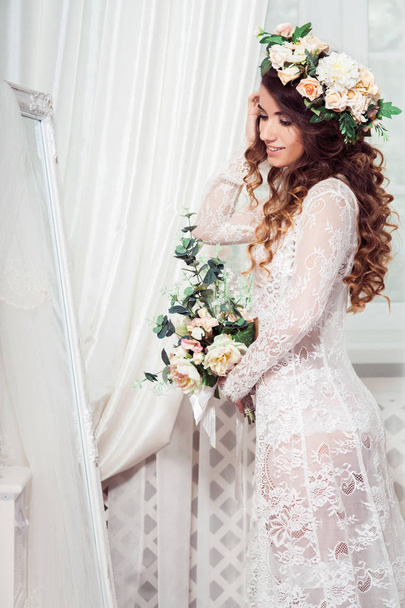 sexy mariée caucasienne debout devant le miroir pleine longueur, vêtue de nuisette blanche, couronne, avec bouquet de fleurs, verticale
 - Photo, image
