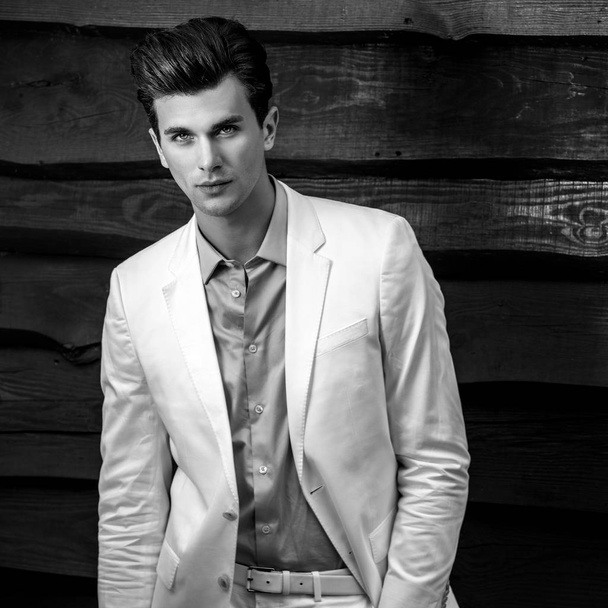 Black-white portrait of young handsome fashionable man in white suit against wooden wall - Foto, Imagen