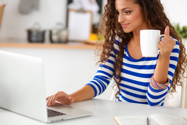 Smiling young woman with coffee cup and laptop in the kitchen at home - Фото, зображення
