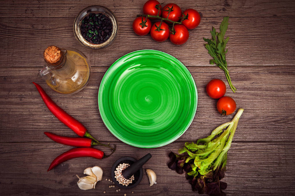 Fresh tomatoes, chili pepper and other spices and herbs around modern green plate in the center of wooden table and cloth napkin. Top view. Blank place for your text. Close-up. - Photo, Image